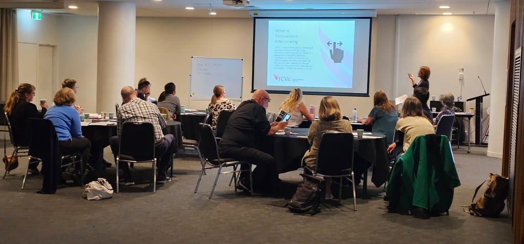 : a photograph from the back of the training room. Attendees are sitting at tables and Robyn Angus is presenting on a screen. The content on screen is titled ‘What is Motivational Interviewing’