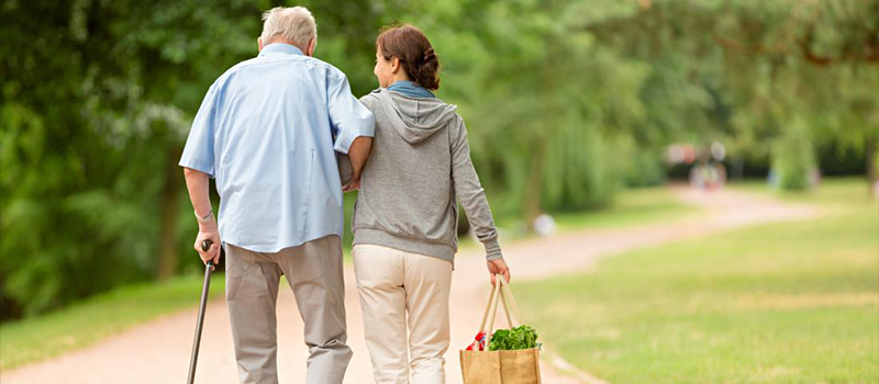 A photograph shows a younger person assisting an older person to walk. The younger person is supporting them and holding a bag of groceries in their other hand.