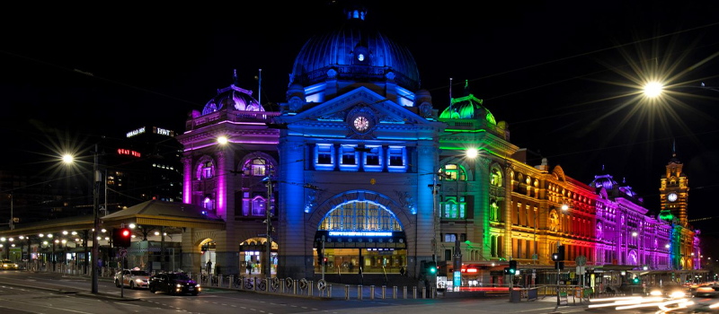 A photo of Flinders Street Station at night with rainbow lights illuminating the facade