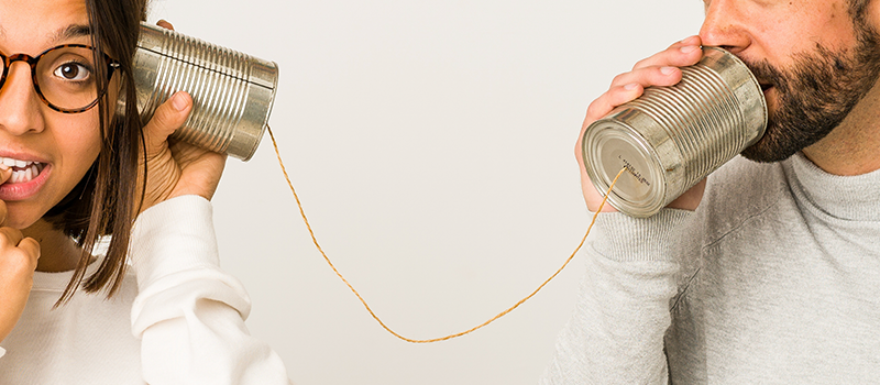 A close up photograph shows two people trying to communicate through tin-can phones. The person listening seems to be confused.