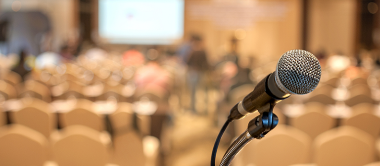 A photograph of a microphone. The background is blurry but can be recognised as a chairs in an auditorium or similar.