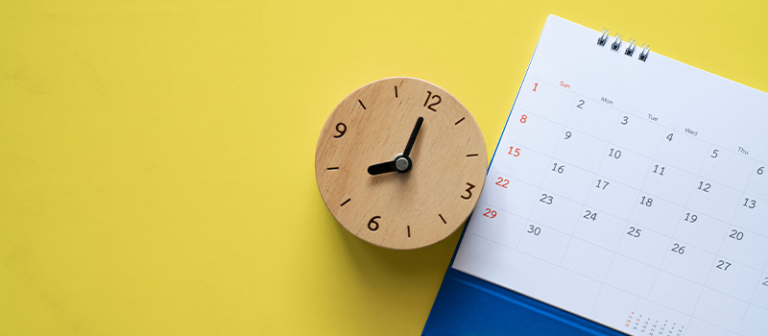 A photograph of a wooden clock next to a calendar, on a bright yellow background.