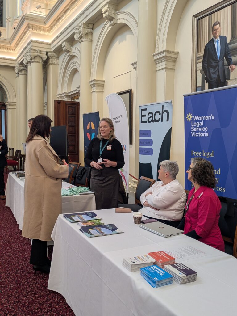 Ann-Marie Hermans MP stands in conversation with a representative at an exhibitor table in the historic Queen's Hall, Parliament House Victoria, with pull-up banners for Gambler's Help, Each and Women's Legal Service Victoria behind, and two seated attendees nearby.