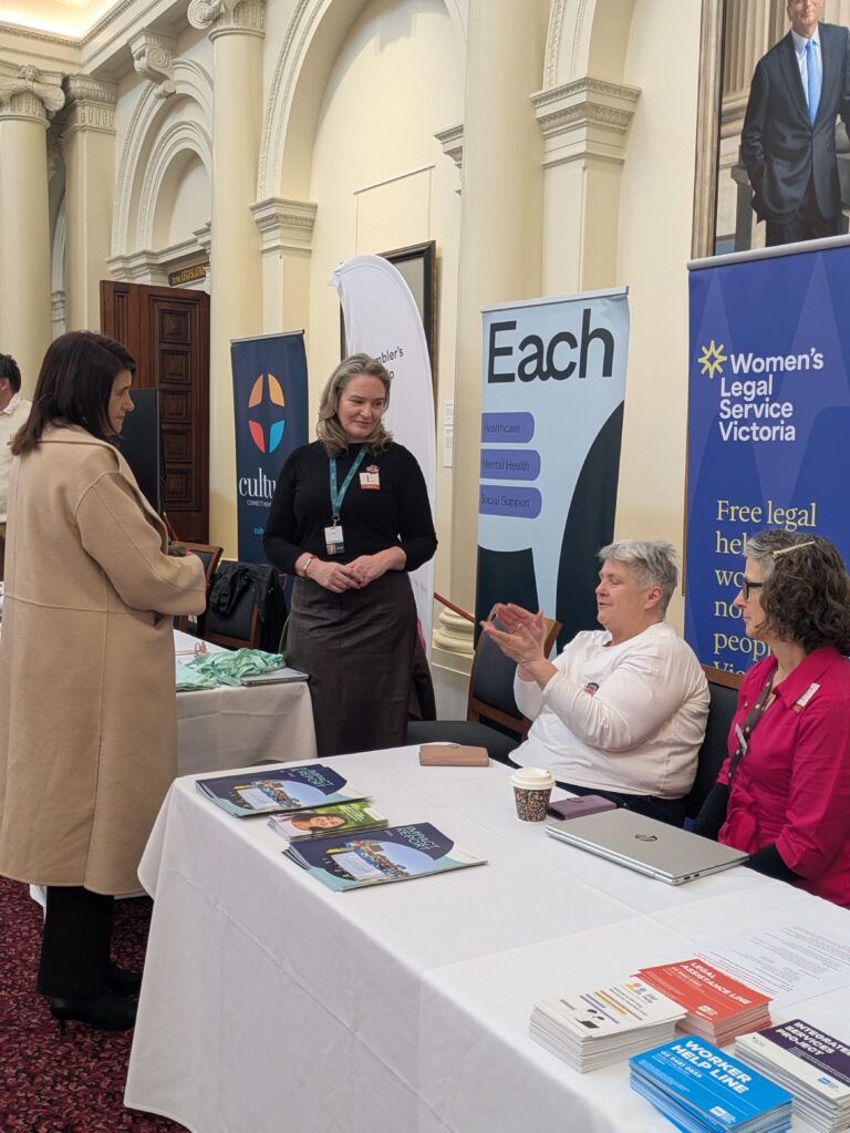 Ann-Marie Hermans MP continues a conversation at the same exhibitor table in Queen's Hall, with a seated attendee applauding and community service banners visible in the background.