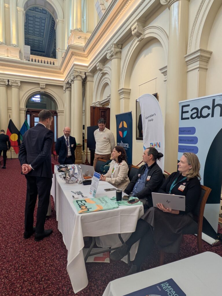 Iwan Walters MP stands with their back to the camera, speaking with three seated attendees at an exhibitor table beneath Queen's Hall's ornate arches, with Aboriginal and Torres Strait Islander flags visible at left.