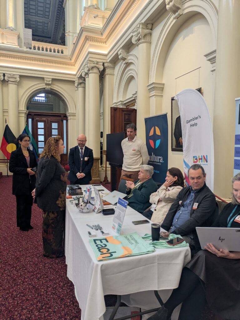 The Hon. Melissa Horne speaks with a small group of attendees and exhibitors gathered around a table in Queen's Hall, with Cultura and Gambler's Help banners in the background.