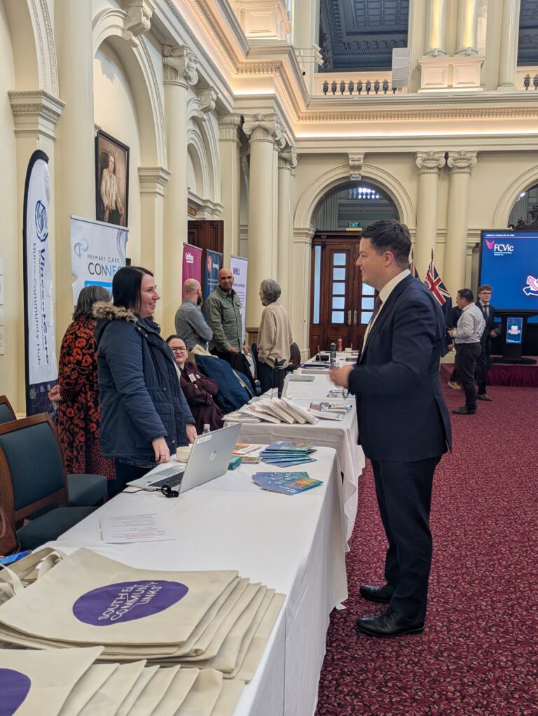 Michael Galea MP speaks with two attendees at an exhibitor table lined with tote bags and information booklets.
