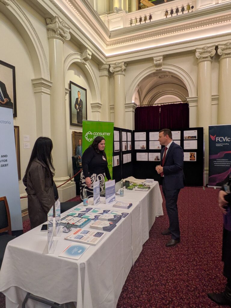 The Hon. Tim Richardson MP speaks with a representative at the Consumer Action Law Centre booth, with the FCVic banner and case study wall in the background.