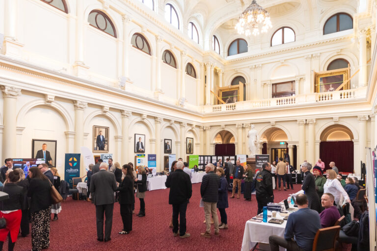 Exhibition booths and attendees filling Queens Hall at Parliament House during the Small Sector, Big Impact showcase, with agency banners including Cultura, EACH and Consumer Action Law Centre.