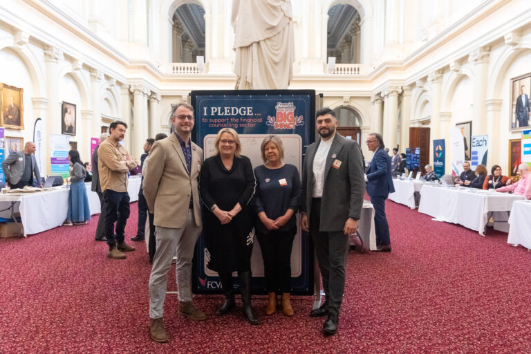 Four people pose in front of the "I pledge to support the financial counselling sector" wall in Queens Hall, including Victorian MP Ros Spence.