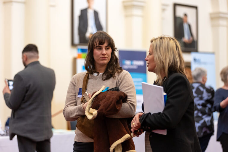 Two attendees in conversation among the exhibition booths in Queens Hall.