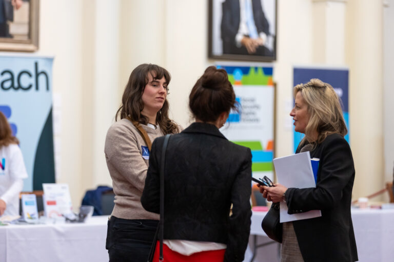 Three attendees in conversation near exhibition banners in Queens Hall.