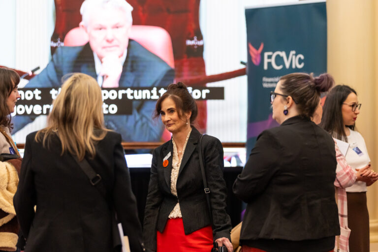 A group of attendees in conversation in front of the FCVic banner and a screen showing parliamentary proceedings.