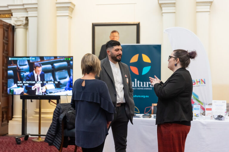 Two attendees speak with a representative from Cultura at their exhibition booth, with parliamentary proceedings showing on a nearby screen.