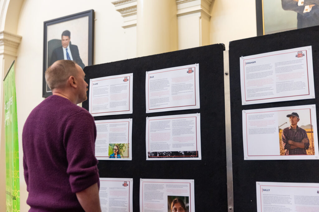 A visitor in a purple jumper reads case study boards, with portraits and a green exhibition banner in the background.