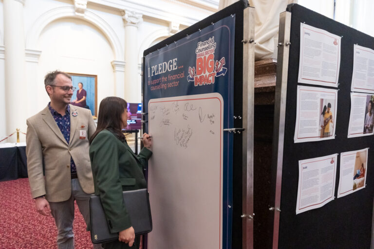 Rachel Payne MP signs the pledge wall while an FCVic representative looks on, with case study panels visible nearby.
