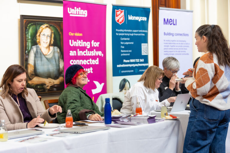 Attendees share food and conversation near the Uniting, Moneycare and MELI exhibition banners.