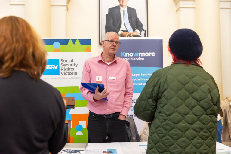 A man speaks with two attendees near the Social Security Rights Victoria and Knowmore Legal Service booths.