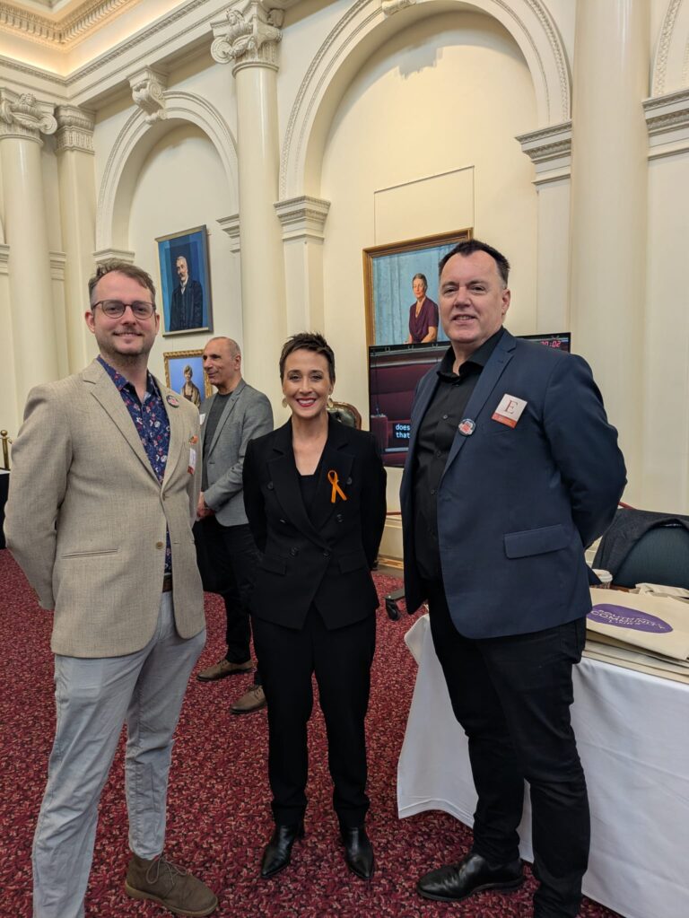 The Hon. Gabrielle Williams, wearing an orange ribbon, stands smiling between two attendees in Queen's Hall, with portrait paintings on the wall behind them.