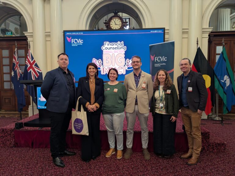 Ann-Marie Hermans MP joins a group of five attendees for a photo in front of the Financial Counselling Small Sector, Big Impact screen, with Australian, Victorian and First Nations flags displayed behind them.