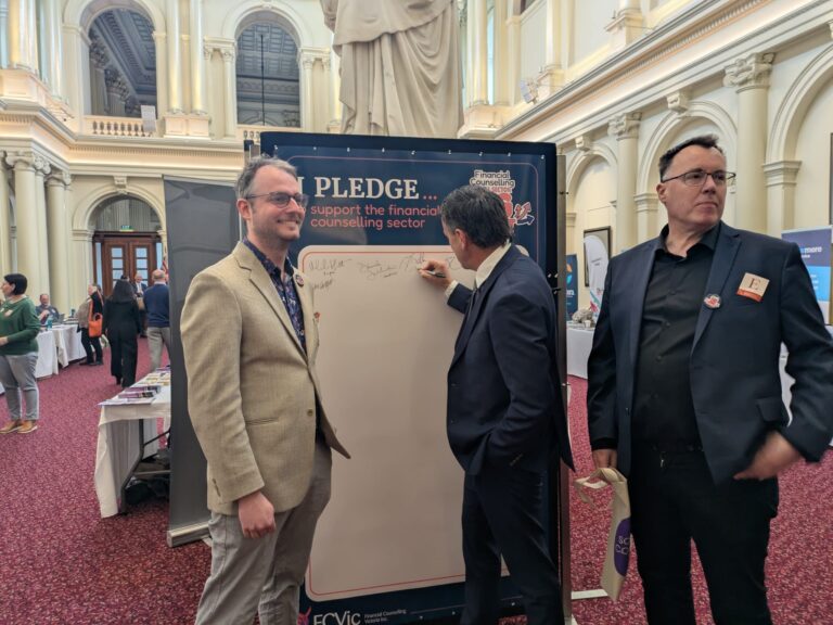 Brad Battin MP signs the "I Pledge" board supporting the financial counselling sector, watched by two attendees in Queen's Hall.