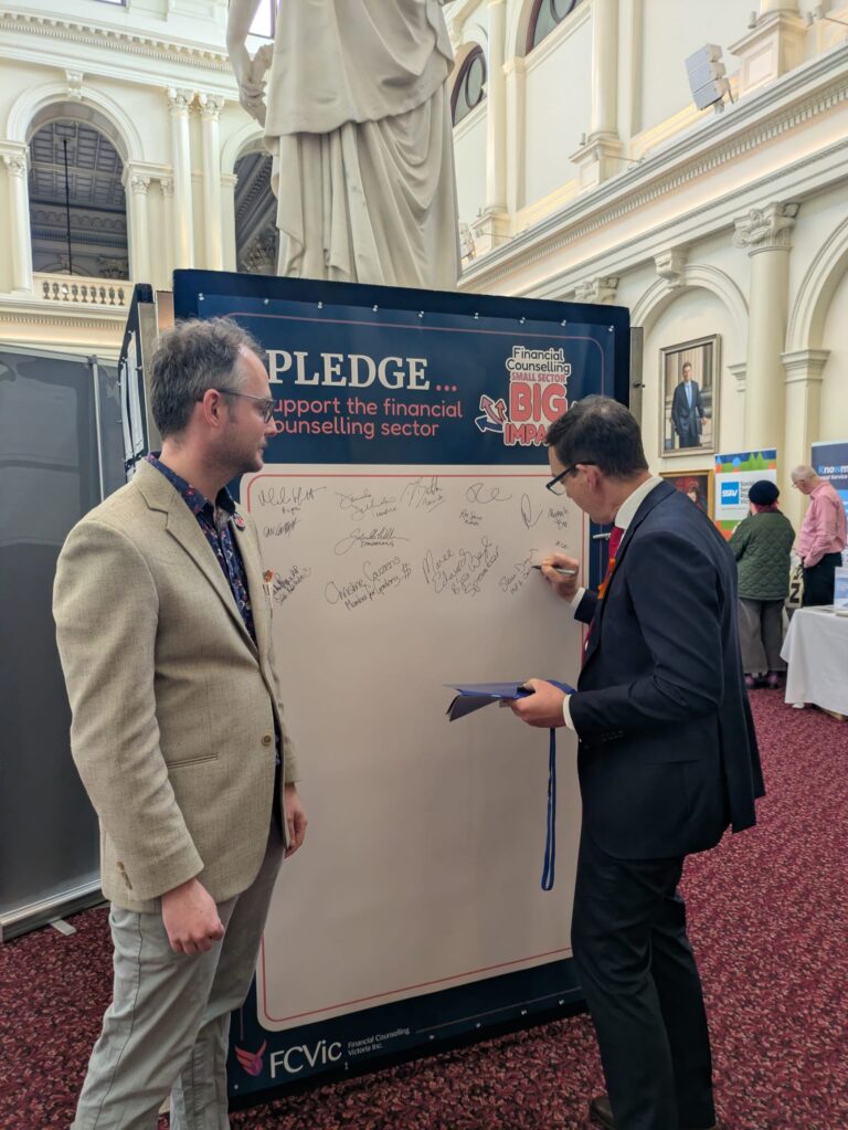 The Hon. Steve Dimopoulos signs the "I Pledge" board supporting the financial counselling sector, with an attendee looking on and other exhibitor stands visible behind.