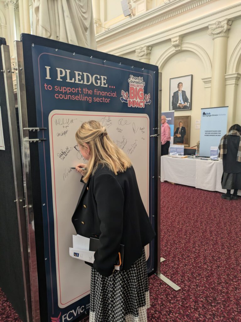 The Hon. Natalie Hutchins signs the "I Pledge" board in Queen's Hall, with exhibitor stands and portraits visible in the background.