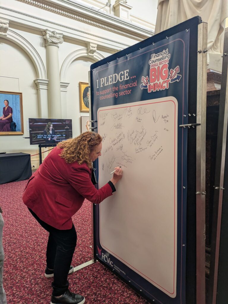 The Hon. Melissa Horne bends to sign the "I Pledge" board, already covered in signatures, beneath a large marble statue in Queen's Hall.