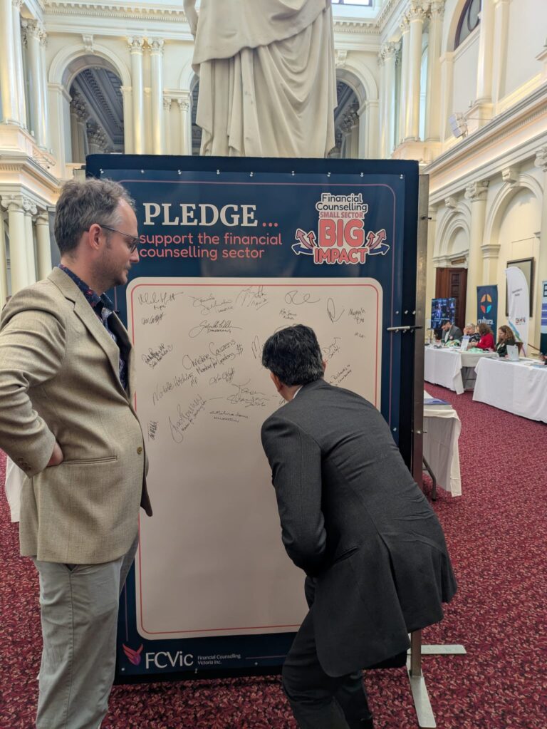 Gary Maas MP leans in to sign the "I Pledge" board, watched by an attendee, with exhibitor tables visible in the background.
