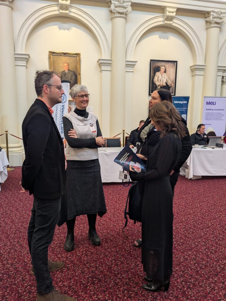Kat Theophanous MP talks with a group of attendees beside exhibitor banners for Moneycare and MeLI in Queen's Hall.