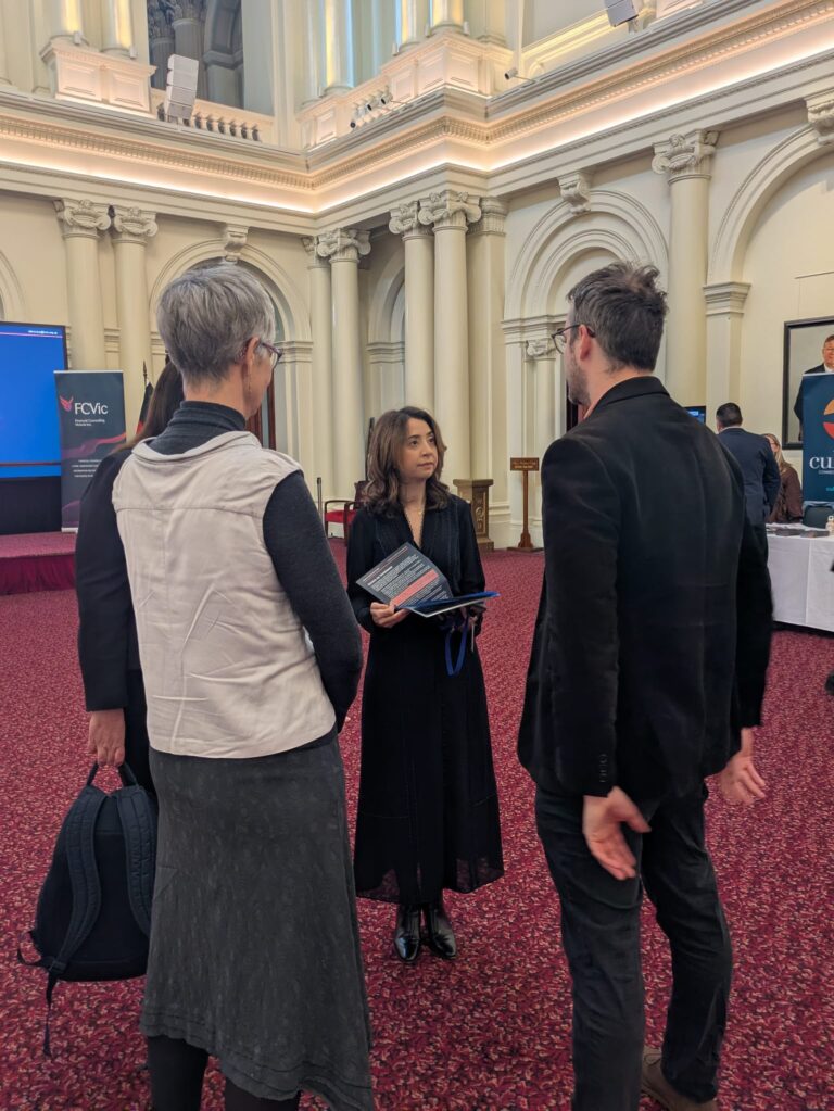 Kat Theophanous MP talks with two attendees in Queen's Hall, Parliament House Victoria, with FCVic and Cultura banners visible behind.