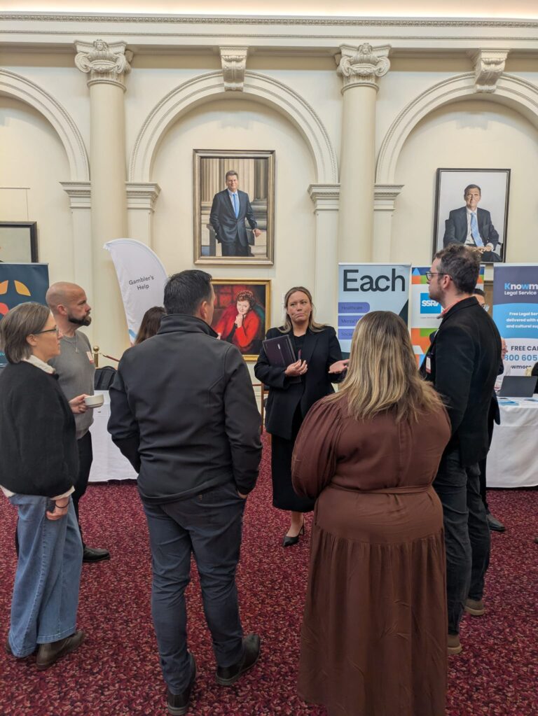 Ella George MP speaks with a group of attendees beneath historic portraits in Queen's Hall, with exhibitor banners for Each, SSRV and Knowmore Legal Service visible behind.