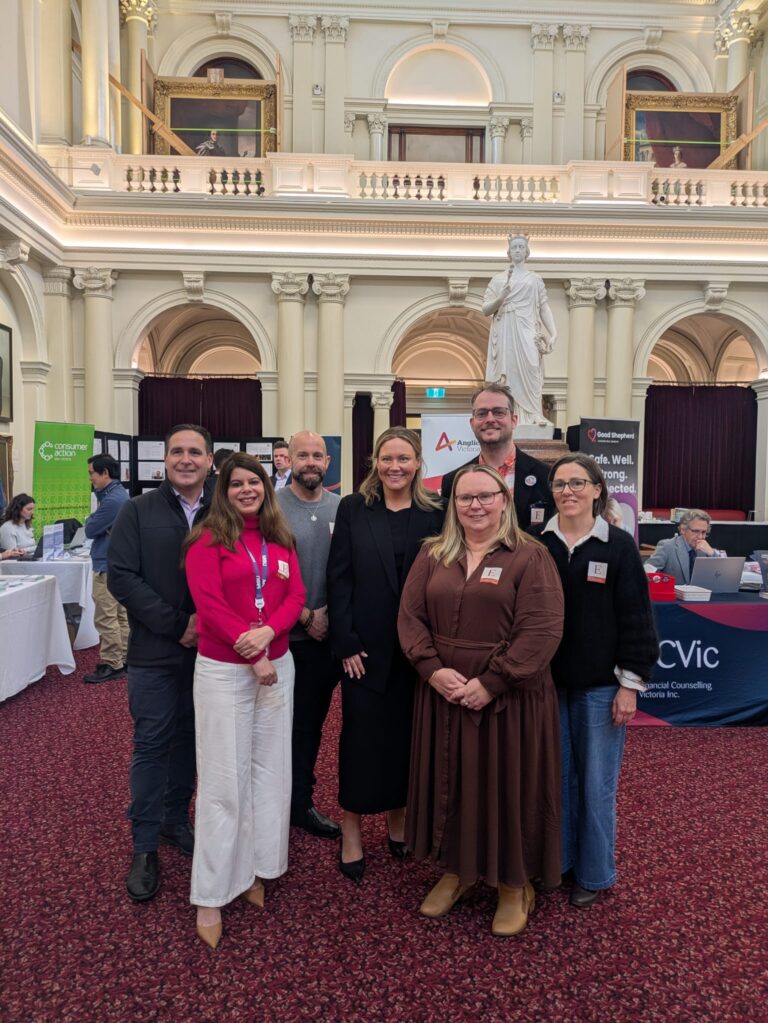 Ella George MP poses with six attendees in front of a marble statue in Queen's Hall, with banners for Consumer Action Law Centre, Anglicare Victoria, Good Shepherd and FCVic visible behind.
