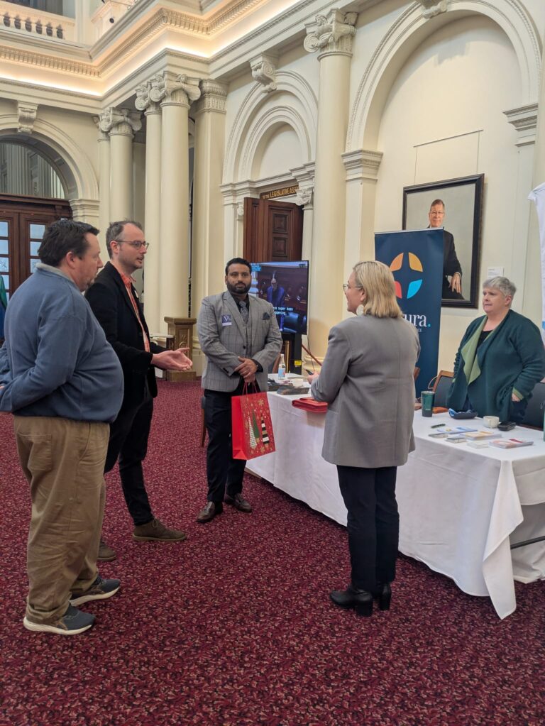 Kathleen Matthews-Ward MP talks with a group of attendees beside a Cultura exhibitor table in Queen's Hall.