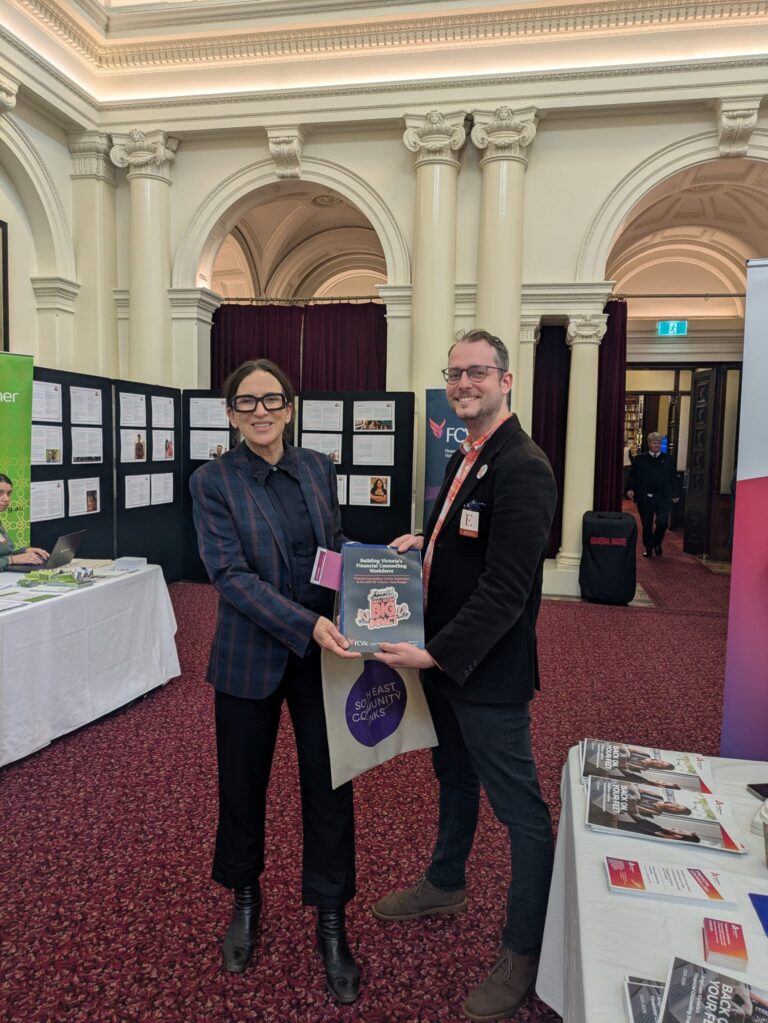 Jordan Crugnale MP stands with an attendee holding a copy of "Building Victoria's Financial Counselling Workforce" in Queen's Hall.