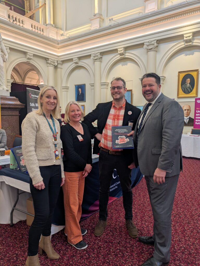 Will Fowles MP poses with three attendees holding a copy of "Building Victoria's Financial Counselling Workforce" in Queen's Hall.