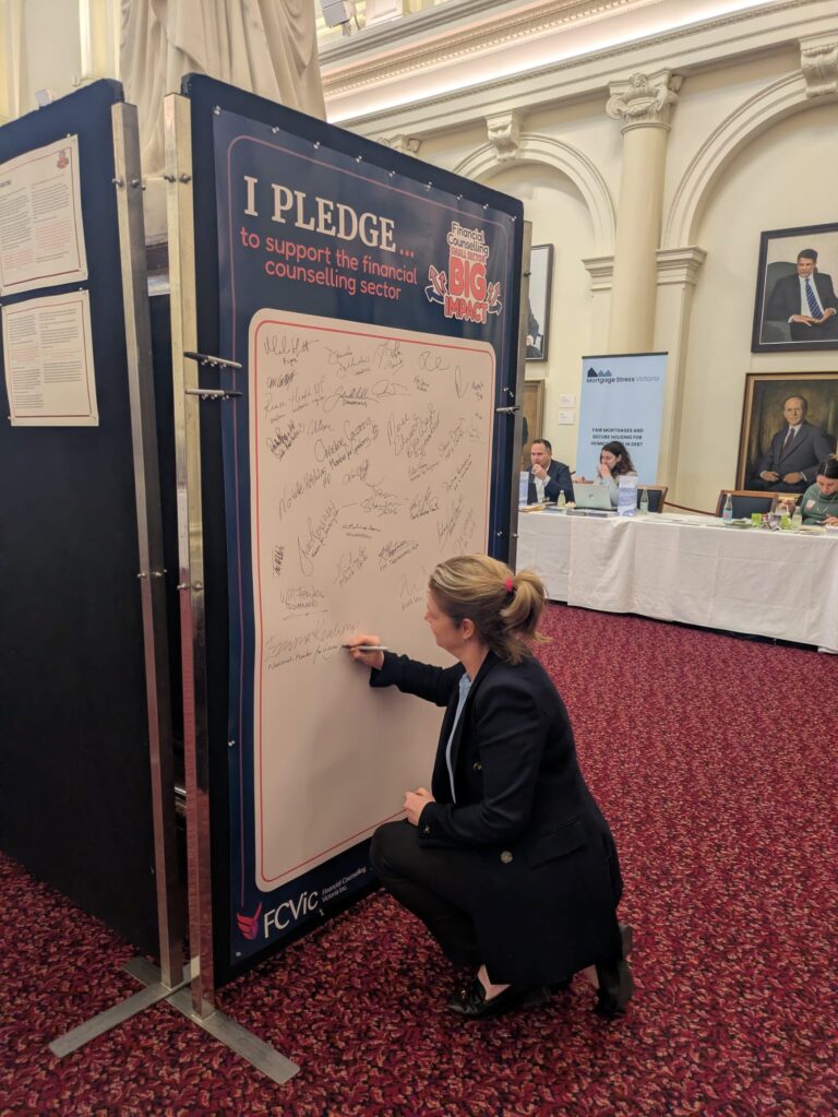 Emma Kealy MP kneels to sign the "I Pledge" board in Queen's Hall, with exhibitor tables visible in the background.