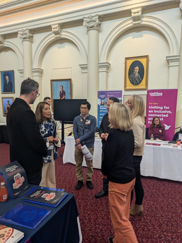 Rachel Westaway MP talks with a group of attendees beside exhibitor banners for South East Community Links and Uniting in Queen's Hall.