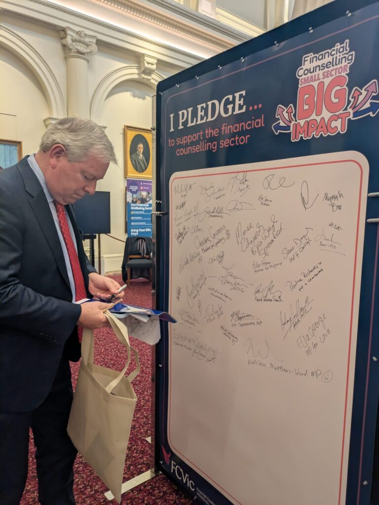 The Hon. David Davis signs the "I Pledge" board in Queen's Hall, with exhibitor banners visible in the background.