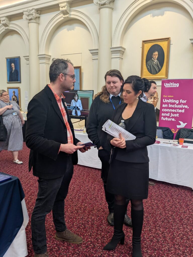 FCVic CEO Zyl Hovenga-Wauchope and an attendee speak with Moira Deeming MP in Queen's Hall, Parliament House Victoria, with a Uniting exhibitor banner visible behind.