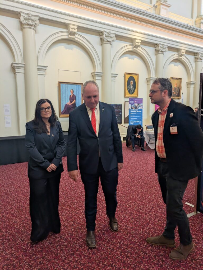 The Hon. Barnaby Joyce MP walks with an attendee and FCVic's CEO through Queen's Hall, past exhibitor banners for South East Community Links.