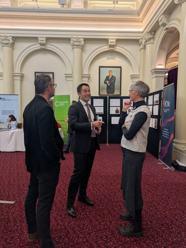 FCVic's CEO speaks with Nathan Lambert MP and an attendee beside display boards in Queen's Hall, with banners for Mortgage Stress Victoria and Consumer Action Law Centre visible behind.