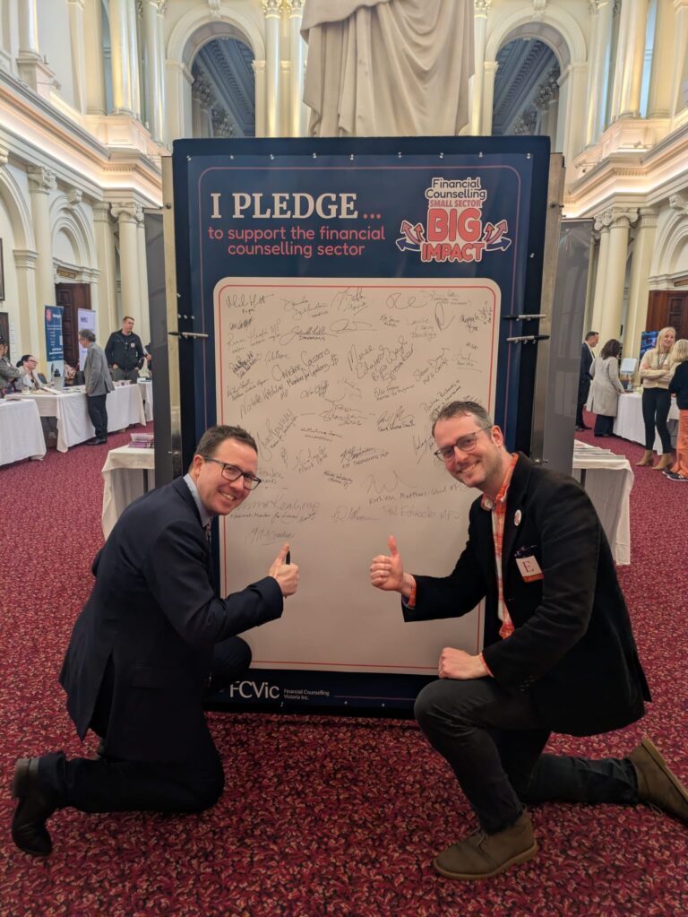 Joe McCracken MP and FCVic's CEO kneel and give a thumbs up beside the "I Pledge" board in Queen's Hall.