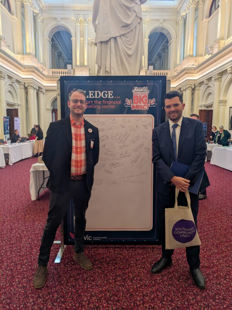 FCVic's CEO stands with Evan Mulholland MP at the "I Pledge" board in Queen's Hall, with an attendee holding a South East Community Links tote bag.