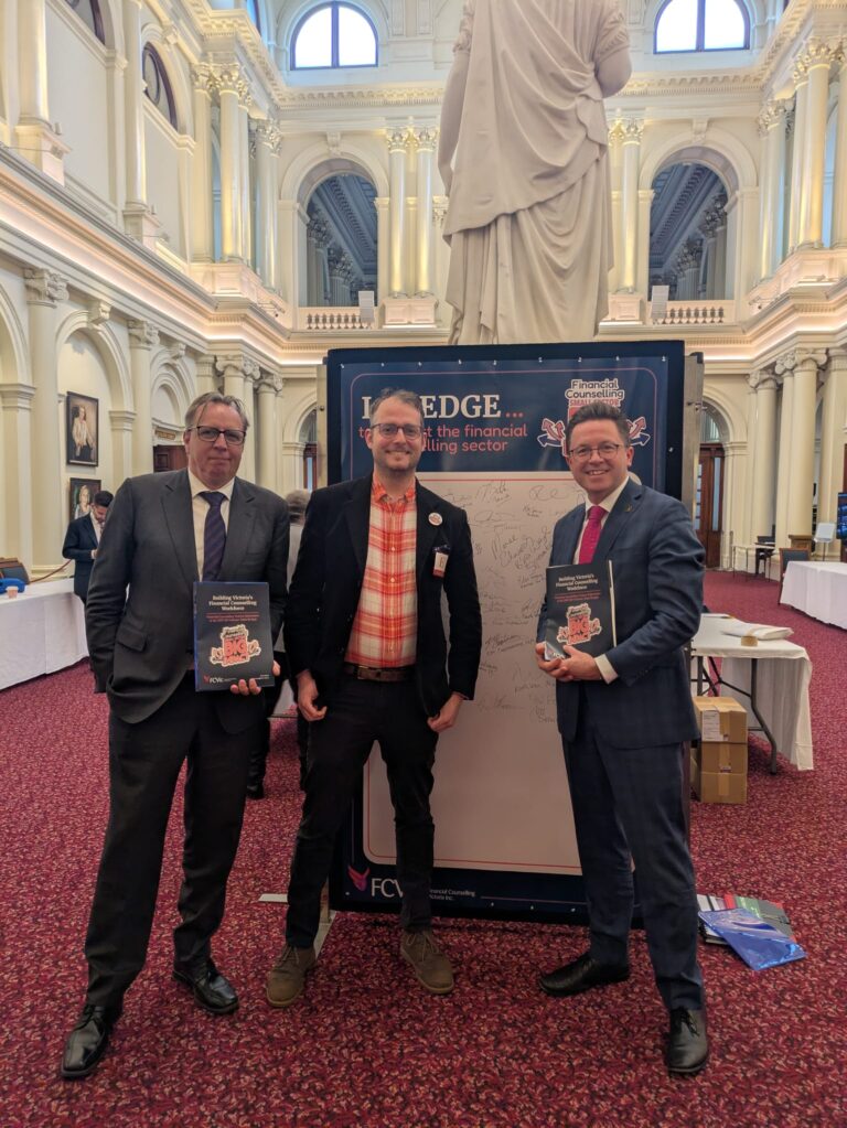 FCVic's CEO stands between The Hon. Anthony Carbines and Matt Fregon MP, all holding copies of "Building Victoria's Financial Counselling Workforce" in Queen's Hall.