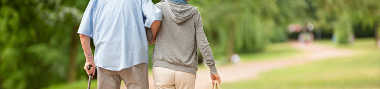An older man is helped by a younger person, holding his arm and carrying his shopping